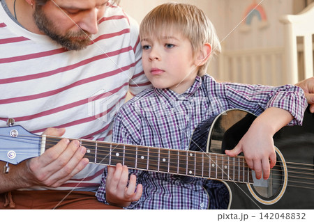 Handsome dad teaches his cute son to play the guitar sitting on sofa in the living room at home. Happy Father's Day. Father teaching his child to play guitar. Kid Learning music skill. Art education 142048832