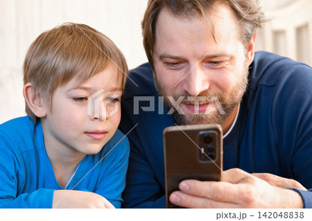 Father and son little boy laughing and using phone. They watching funny videos together, playing games on smartphone. Concept technology, smart phone addicted. Fun weekend activity. Modern fatherhood Father and son little boy laughing and using phone. They watching funny videos together, playing games on smartphone. Concept technology, smart phone addicted. Fun weekend activity. Modern fatherhood 142048838