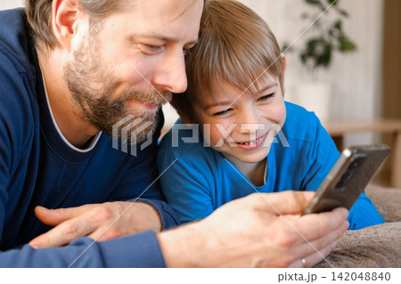 Father and son little boy laughing and using phone. They watching funny videos together, playing games on smartphone. Concept technology, smart phone addicted. Fun weekend activity. Modern fatherhood Father and son little boy laughing and using phone. They watching funny videos together, playing games on smartphone. Concept technology, smart phone addicted. Fun weekend activity. Modern fatherhood 142048840