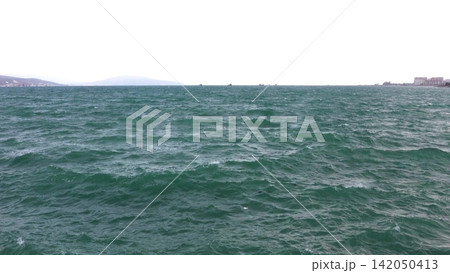 Stormy sea with powerful waves, distant city skyline and ships on the horizon under dramatic cloudy sky Stormy sea with powerful waves, distant city skyline and ships on the horizon under dramatic cloudy sky 142050413