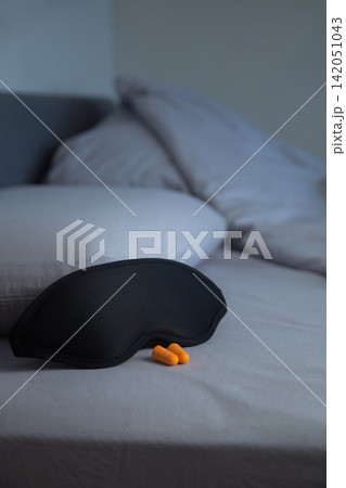 On the light gray bed linen, against the background of a pillow, there is a black sleep mask, next to which there are two bright orange earplugs. Concept: a complete set for complete isolation from On the light gray bed linen, against the background of a pillow, there is a black sleep mask, next to which there are two bright orange earplugs. Concept: a complete set for complete isolation from 142051043