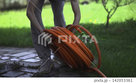 Person watering a backyard path with a coiled orange hose Person watering a backyard path with a coiled orange hose 142053361