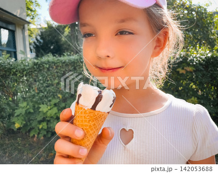 Cheerful girl holding vanilla chocolate ice cream cone in garden. Happy child smiles while enjoying a refreshing summer dessert outdoors. Childhood joy concept. Cheerful girl holding vanilla chocolate ice cream cone in garden. Happy child smiles while enjoying a refreshing summer dessert outdoors. Childhood joy concept. 142053688