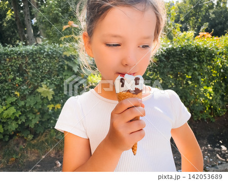 Little girl licking vanilla chocolate ice cream in sunny garden. Happy child enjoys a creamy frozen dessert outdoors. Summer treat concept. Little girl licking vanilla chocolate ice cream in sunny garden. Happy child enjoys a creamy frozen dessert outdoors. Summer treat concept. 142053689
