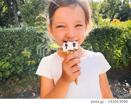 Cheerful girl smiling while eating vanilla chocolate ice cream outdoors. Happy child enjoys a sweet frozen dessert in sunlight. Childhood joy concept. Cheerful girl smiling while eating vanilla chocolate ice cream outdoors. Happy child enjoys a sweet frozen dessert in sunlight. Childhood joy concept. 142053690