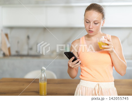 Teenage girl looking at smartphone while drinking fresh juice in kitchen Teenage girl looking at smartphone while drinking fresh juice in kitchen 142055745