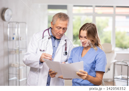 Middle-aged nurse and mature doctor discussing diagnosis standing in cabinet Middle-aged nurse and mature doctor discussing diagnosis standing in cabinet 142056352