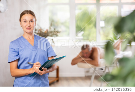 Positive middle-aged masseuse posing with folder in hand in the therapy room Positive middle-aged masseuse posing with folder in hand in the therapy room 142056356