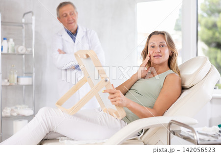 Happy woman examining her face after cosmetology procedures in clinic Happy woman examining her face after cosmetology procedures in clinic 142056523