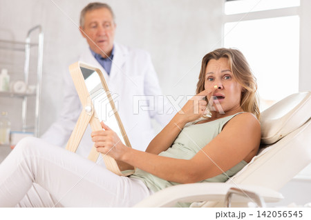 Unsatisfied middle-aged woman holding mirror lying on clinical chair Unsatisfied middle-aged woman holding mirror lying on clinical chair 142056545