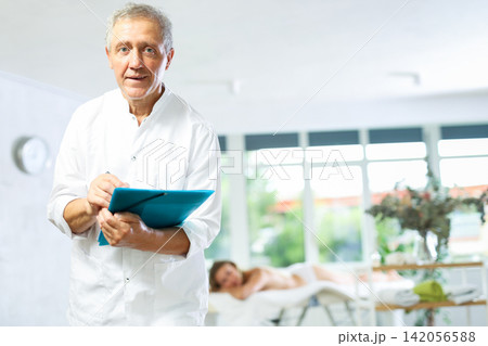 Positive old masseur posing with folder in hand in the therapy room 142056588