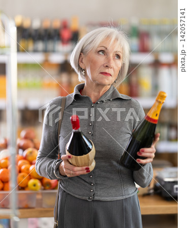 Shopping in produce supermarket - mature female buyer makes choice bottle of wine 142057141