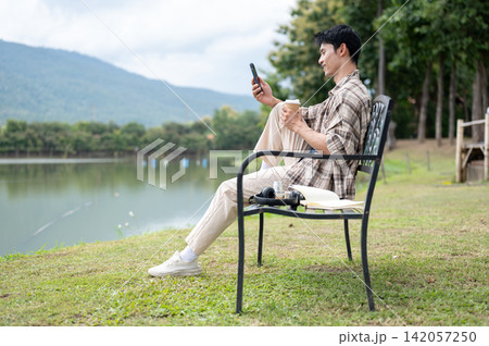 Young Asian man relaxing with phone and coffee on a bench by a scenic mountain lake in greenery park Young Asian man relaxing with phone and coffee on a bench by a scenic mountain lake in greenery park 142057250