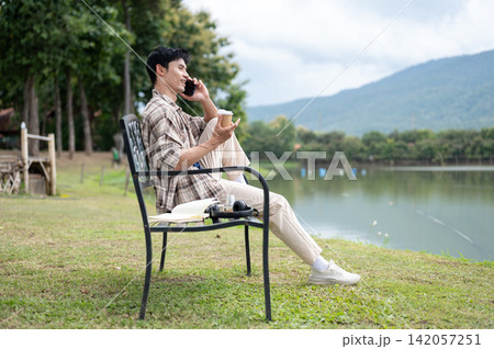 Young Asian man talking on phone while enjoying a coffee on bench at lakeside surrounded by mountain Young Asian man talking on phone while enjoying a coffee on bench at lakeside surrounded by mountain 142057251
