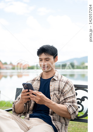 Young Asian man sitting comfortably while checking his smartphone on bench by the lake in the park. 142057264