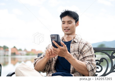 Asian Man Sitting on Bench and Smiling while Browsing Smartphone Asian Man Sitting on Bench and Smiling while Browsing Smartphone 142057265