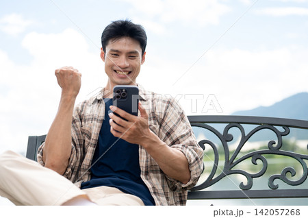 Young Asian man making a fist in excited as looking at smartphone on bench in park under clear sky. Young Asian man making a fist in excited as looking at smartphone on bench in park under clear sky. 142057268