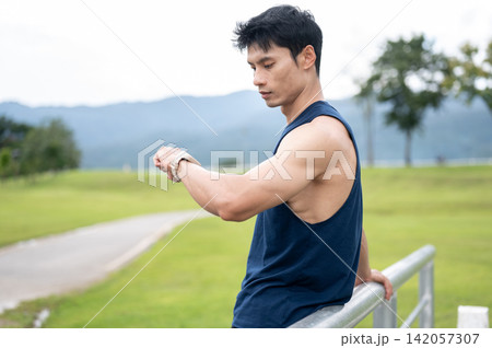 Athletic Asian man checking time on wristwatch after exercising in a green field park on sunny day. 142057307