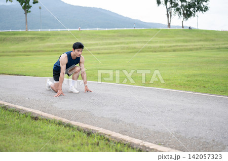 Athletic Asian man sitting crouching preparing to run on the road along a wide green park landscape. Athletic Asian man sitting crouching preparing to run on the road along a wide green park landscape. 142057323