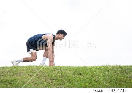 Athletic Asian man crouching preparing to run on the road over a grassy field under clear bright sky 142057335