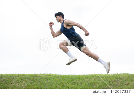 Athletic Asian man in running posture floating over a road along grass field under clear bright sky. 142057337