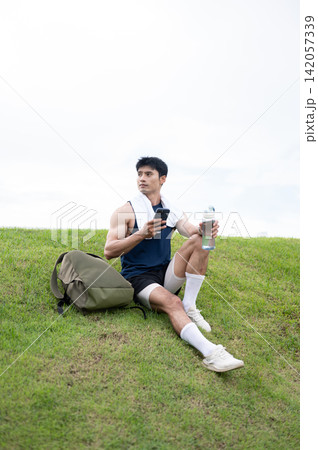 Athletic Asian man resting with phone and water bottle on grass after exercising in the green park. Athletic Asian man resting with phone and water bottle on grass after exercising in the green park. 142057339