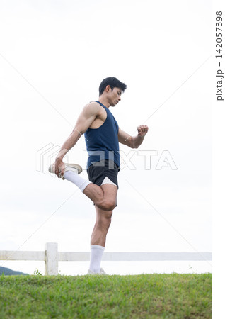 Full body of athletic asian man stretching legs over a grassy field in green park under a bright sky 142057398