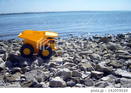 A bright yellow toy dump truck rests on a shore covered with grey and brown rocks. In the background, the wide blue water stretches to the horizon under a clear sky on a sunny day A bright yellow toy dump truck rests on a shore covered with grey and brown rocks. In the background, the wide blue water stretches to the horizon under a clear sky on a sunny day 142059584