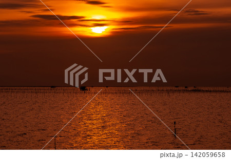 Orange sunset sky illuminating tranquil coastal fishing village with wooden huts, aquaculture structures and shimmering sea reflections, capturing a serene tropical landscape at golden hour. Orange sunset sky illuminating tranquil coastal fishing village with wooden huts, aquaculture structures and shimmering sea reflections, capturing a serene tropical landscape at golden hour. 142059658