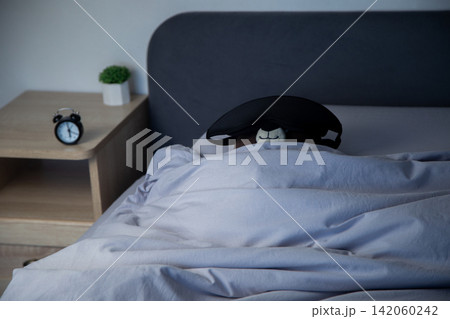 Top view on a gray bed under a blanket lies a brown teddy bear with a black sleep mask, next to a bedside table there is a mini alarm clock and a white pot with a plant. The concept of a sleeping Top view on a gray bed under a blanket lies a brown teddy bear with a black sleep mask, next to a bedside table there is a mini alarm clock and a white pot with a plant. The concept of a sleeping 142060242