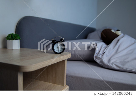 There is a mini alarm clock and a white potted plant on the bedside table, and a brown teddy bear is lying on a gray bed under a blanket in the background. The concept of a sleep mode, a comfortable There is a mini alarm clock and a white potted plant on the bedside table, and a brown teddy bear is lying on a gray bed under a blanket in the background. The concept of a sleep mode, a comfortable 142060243