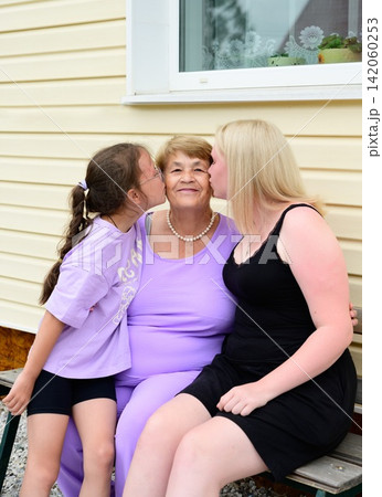 Two granddaughters kissing happy Russian grandmother showing genuine connection at rural veranda. Two granddaughters kissing happy Russian grandmother showing genuine connection at rural veranda. 142060253