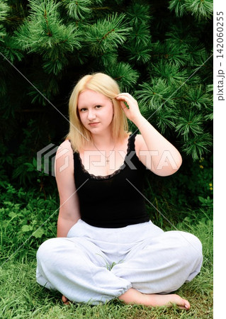 17 years old Russian blonde teenager girl sitting cross legged against lush green pine tree. 17 years old Russian blonde teenager girl sitting cross legged against lush green pine tree. 142060255