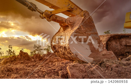 Dirt metal bucket of backhoe after digging soil. Backhoe parked at agricultural land on sunset sky background. Crawler excavator. Earthmoving machine at construction site at dusk. Excavation vehicle. Dirt metal bucket of backhoe after digging soil. Backhoe parked at agricultural land on sunset sky background. Crawler excavator. Earthmoving machine at construction site at dusk. Excavation vehicle. 142060938