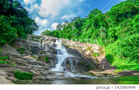 Beautiful tropical waterfall cascading over granite mountain rocks beneath blue sky. Lush rainforest in Thailand. Wilderness landscape for travel and nature theme. Ecotourism and conservation. Beautiful tropical waterfall cascading over granite mountain rocks beneath blue sky. Lush rainforest in Thailand. Wilderness landscape for travel and nature theme. Ecotourism and conservation. 142060994