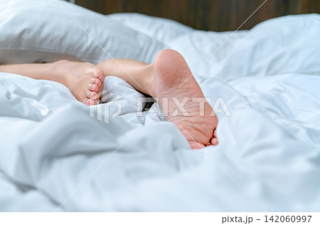 Woman barefoot on bed over white linen blanket and bed sheet in hotel or home bedroom. Sleep and relaxation. Lazy Sunday morning. Bare feet of woman chilling sleep on white comfort bed and duvet. Woman barefoot on bed over white linen blanket and bed sheet in hotel or home bedroom. Sleep and relaxation. Lazy Sunday morning. Bare feet of woman chilling sleep on white comfort bed and duvet. 142060997