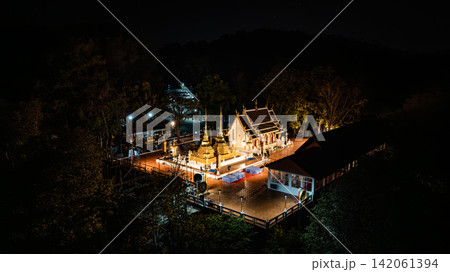 Phra That Doi Tung rises above the dark highland landscape, its radiant golden architecture contrasting with the forest and distant lights in a serene aerial scene from 2023. Phra That Doi Tung rises above the dark highland landscape, its radiant golden architecture contrasting with the forest and distant lights in a serene aerial scene from 2023. 142061394