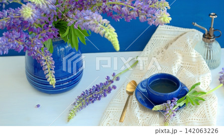 Purple lupines in a blue ceramic vase and a blue cup of tea on a blue background. Lifestyle, still life Purple lupines in a blue ceramic vase and a blue cup of tea on a blue background. Lifestyle, still life 142063226