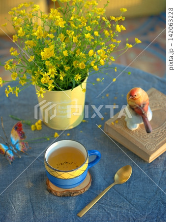 A ceramic blue cup with tea and yellow wildflowers. A blue and light blue color palette. A ceramic blue cup with tea and yellow wildflowers. A blue and light blue color palette. 142063228