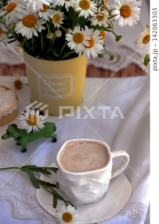 Still life with cappuccino in a white cup, cakes and daisies on a white tablecloth Still life with cappuccino in a white cup, cakes and daisies on a white tablecloth 142063303