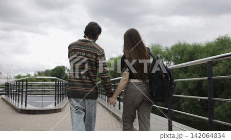 Young people walking hand in hand across metal-railed pedestrian bridge under overcast sky, rear view with casual clothing, calm outdoor urban setting, soft natural light and muted colors Young people walking hand in hand across metal-railed pedestrian bridge under overcast sky, rear view with casual clothing, calm outdoor urban setting, soft natural light and muted colors 142064834