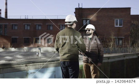 Construction workers shaking hands near red brick industrial building beside water pool, side view under daylight, showing professional agreement, conversation, teamwork, and site collaboration Construction workers shaking hands near red brick industrial building beside water pool, side view under daylight, showing professional agreement, conversation, teamwork, and site collaboration 142064874