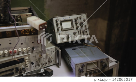 Vintage analog oscilloscopes and test instruments stacked on cluttered workshop bench under warm directional light, with worn cables, knobs, metal housings, and soft shadowed detail 142065017