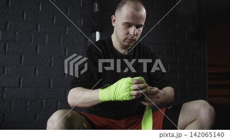 Male athlete wrapping neon green hand wraps before boxing workout inside dim gym with dark brick wall, focused posture, strong contrast, and gritty preparation mood Male athlete wrapping neon green hand wraps before boxing workout inside dim gym with dark brick wall, focused posture, strong contrast, and gritty preparation mood 142065140