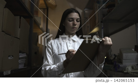 Young woman reviewing inventory notes in a narrow storage room Young woman reviewing inventory notes in a narrow storage room 142065153