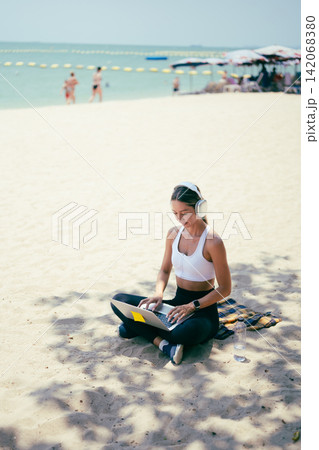 Side view of fit woman using laptop while sitting on the sand at a tropical beach 142068380