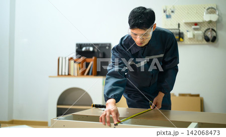 Asian Male Technician in Uniform Measuring Furniture Panel with Tape Measure in Workshop Asian Male Technician in Uniform Measuring Furniture Panel with Tape Measure in Workshop 142068473