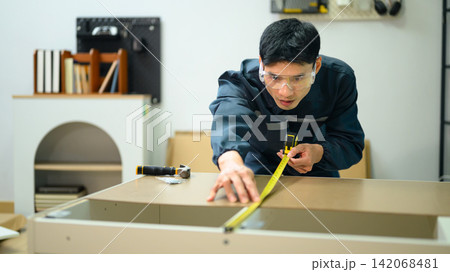Asian Male Technician in Uniform Measuring Furniture Panel with Tape Measure in Workshop Asian Male Technician in Uniform Measuring Furniture Panel with Tape Measure in Workshop 142068481