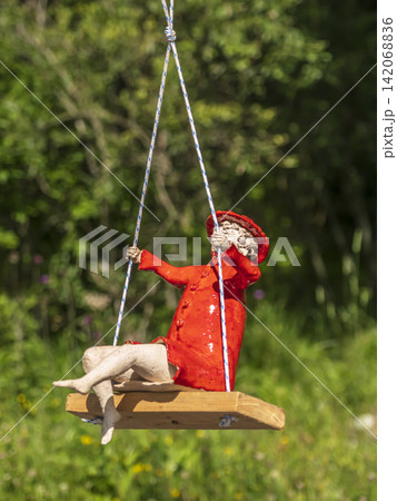 Red-Coated Ceramic Figurine On Wooden Swing In Sunny Garden Landscape With Playful Summer Mood 142068836