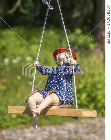Ceramic Doll On Wooden Swing In Garden Wearing Blue Polka Dot Dress And Red Hat 142068837